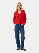 A woman modeling a bright red long-sleeve V-neck sweater paired with dark blue jeans and black flats, standing against a light gray studio background.