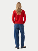 A woman viewed from behind wearing a bright red crewneck sweatshirt and blue jeans, standing against a light gray studio background.