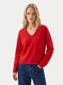A blonde woman wearing a bright red long-sleeve V-neck knit sweater and blue jeans, standing against a light gray studio background.