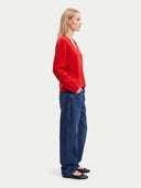 A woman shown in profile wearing a bright red long-sleeve knit top, dark blue wide-leg jeans, and black flats, standing against a plain light gray studio background.