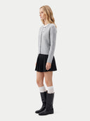 A woman stands in profile wearing a light gray ribbed cardigan over a white collared shirt, a black pleated mini skirt, white knee-high socks, and black knee-high boots, against a plain light gray studio background.