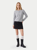 A blonde model stands against a light gray studio background wearing a light gray ribbed button-front cardigan, a black mini skirt, white knee-high socks, and black boots.