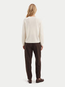 A woman viewed from the back wearing a cream long-sleeve knit sweater and dark brown trousers, standing against a plain light gray studio background.