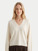 A blonde woman wearing a cream-colored V-neck knit sweater and dark brown trousers, photographed against a solid light gray background.