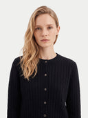 A blonde woman wearing a black cable-knit button-front cardigan sweater, photographed against a clean light gray studio background.