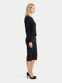 A woman stands in profile wearing a black long-sleeve button-front cardigan, black cropped trousers, and black high heels against a plain light gray studio background.