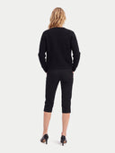 A woman viewed from the back wearing a black ribbed knit sweater, black cropped capri pants, and black pointed-toe heels against a light gray studio background.