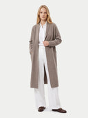 A woman stands against a light gray studio background wearing a long taupe knit cardigan over a white button-up shirt and white wide-leg trousers, with dark brown loafers.