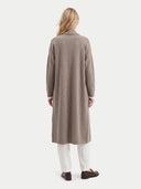 A woman viewed from the back wearing a long taupe knit cardigan over white trousers and brown loafers, photographed against a clean off-white studio background.