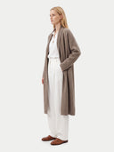 A woman stands in profile wearing a long taupe knit cardigan over a white top and white wide-leg trousers, with brown loafers, against a plain light gray studio background.