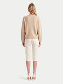 A woman viewed from the back wearing a light beige knit sweater, off-white cropped trousers, and white high-heeled sandals against a plain light gray studio background.