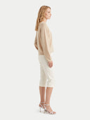 A woman shown in side profile wearing a light beige long-sleeve knit sweater, white cropped pants, and white strappy high-heeled sandals against a plain light gray studio background.