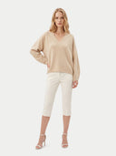 A woman modeling a beige long-sleeve V-neck knit sweater paired with off-white cropped pants and white heeled sandals, photographed against a plain light gray studio background.