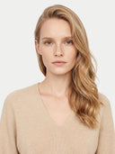 A woman with blonde hair wearing a beige ribbed V-neck sweater, photographed against a clean light gray background.