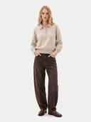 A woman stands against a light gray studio background wearing a beige knit polo sweater with a small embroidered chest logo, dark brown wide-leg corduroy pants, and brown leather shoes.
