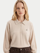 A blonde woman wearing a light beige knit polo sweater with a small circular embroidered logo on the chest, paired with dark brown jeans, against a plain light gray background.