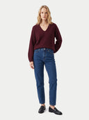 A woman modeling a dark burgundy V-neck knit sweater and blue jeans with black loafers, standing against a light gray studio background.