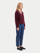 A woman modeling a long-sleeve V-neck knit sweater in deep burgundy, paired with blue jeans and black loafers, against a light gray studio background.