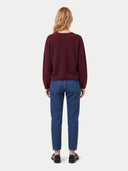 A woman viewed from the back wearing a maroon long-sleeve knit sweater and blue jeans, standing against a light gray studio background.