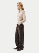 A woman stands in profile wearing a light beige knit quarter-zip pullover, dark brown wide-leg corduroy pants, and dark brown leather shoes against a plain light gray studio background.