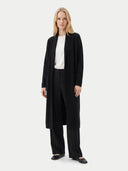 A blonde model stands against a light gray studio background wearing a long black open-front cardigan over a white top and black wide-leg trousers.