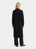 A woman viewed from the back wearing a long black coat, wide-leg black trousers, and black flats against a plain light gray studio background.