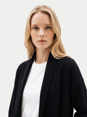 A blonde woman wearing a black open-front cardigan over a white crewneck t-shirt, photographed against a clean, light gray studio background.