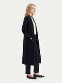 A woman stands in profile wearing a long black open-front cardigan over a white top and black pants, photographed against a plain light gray studio background.