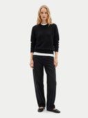A woman stands against a light gray studio background wearing a black crewneck sweatshirt over a white undershirt, black wide-leg pants, and black flats.