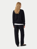 A woman with long blonde hair is shown from the back wearing a black crewneck sweatshirt with a tonal embroidered logo, black pants, and black flats, against a light gray studio background.