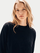 A woman with blonde hair wearing a black crewneck sweatshirt with a tonal embroidered logo on the front, photographed against a light gray studio background.
