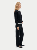 A woman stands in profile wearing a black long-sleeve knit top, black wide-leg trousers, and black flats against a clean light gray studio background.