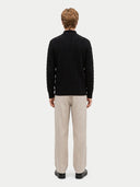 A man viewed from the back wearing a black cable-knit mock-neck sweater and beige trousers, standing against a light gray studio background.