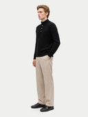A man wearing a black cable-knit mock-neck sweater with a buttoned placket, beige linen trousers, and black leather shoes, standing against a light gray studio background.