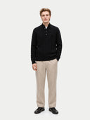 A man modeling a black cable-knit polo sweater with beige trousers and black shoes, standing against a light gray studio background.