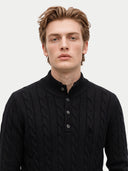 A young man with light brown hair wearing a black cable-knit mock-neck sweater with a three-button placket, photographed against a solid light gray background.
