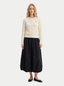 A woman stands against a light gray studio background wearing a cream cable-knit cardigan with a small embroidered logo on the chest, a long black skirt, and black flats.