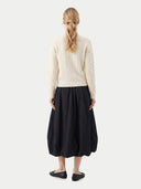 back view of a woman wearing a white cable knit sweater and a black skirt with flat on a light gray background.