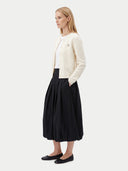 A woman stands in profile wearing a cream cable-knit cardigan over a white top, paired with a black pleated midi skirt and black flats, against a light gray studio background.