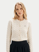 A blonde woman wearing a cream cable-knit cardigan sweater with a small embroidered logo on the chest, paired with a black skirt, against a light gray studio background.