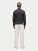 A man viewed from behind wearing a dark charcoal gray mock neck sweater and off-white trousers, standing against a light gray studio background.