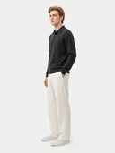 A man wearing a dark charcoal long-sleeve polo shirt, off-white linen trousers, and white sneakers, standing in profile against a light gray studio background.