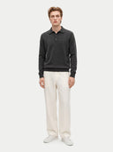 A man modeling a dark charcoal gray knit polo sweater paired with off-white trousers and white sneakers, standing against a solid light gray background.