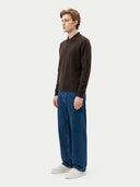 A man stands in profile wearing a dark brown long-sleeve polo sweater, wide-leg blue jeans, and light beige sneakers against a plain light gray studio background.
