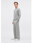 A man modeling a light gray knit crewneck sweater and matching wide-leg sweatpants, paired with white sneakers, against a plain light gray studio background.