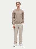 A man modeling a light taupe crewneck sweater and beige trousers with white sneakers, standing against a solid light gray background.
