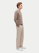 A man in a side profile wearing a taupe crewneck sweater, beige corduroy trousers, and white sneakers, standing against a light gray studio background.
