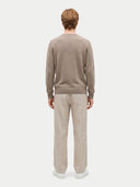 A man viewed from behind wearing a taupe knit crewneck sweater and light beige trousers, standing against a light gray studio background.