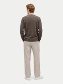 A man viewed from behind wearing a dark brown long-sleeve knit sweater and light beige trousers, standing against a plain light gray studio background.