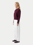 A woman stands in profile wearing a dark burgundy long-sleeve polo sweater, white wide-leg trousers, and dark burgundy loafers against a light gray studio background.